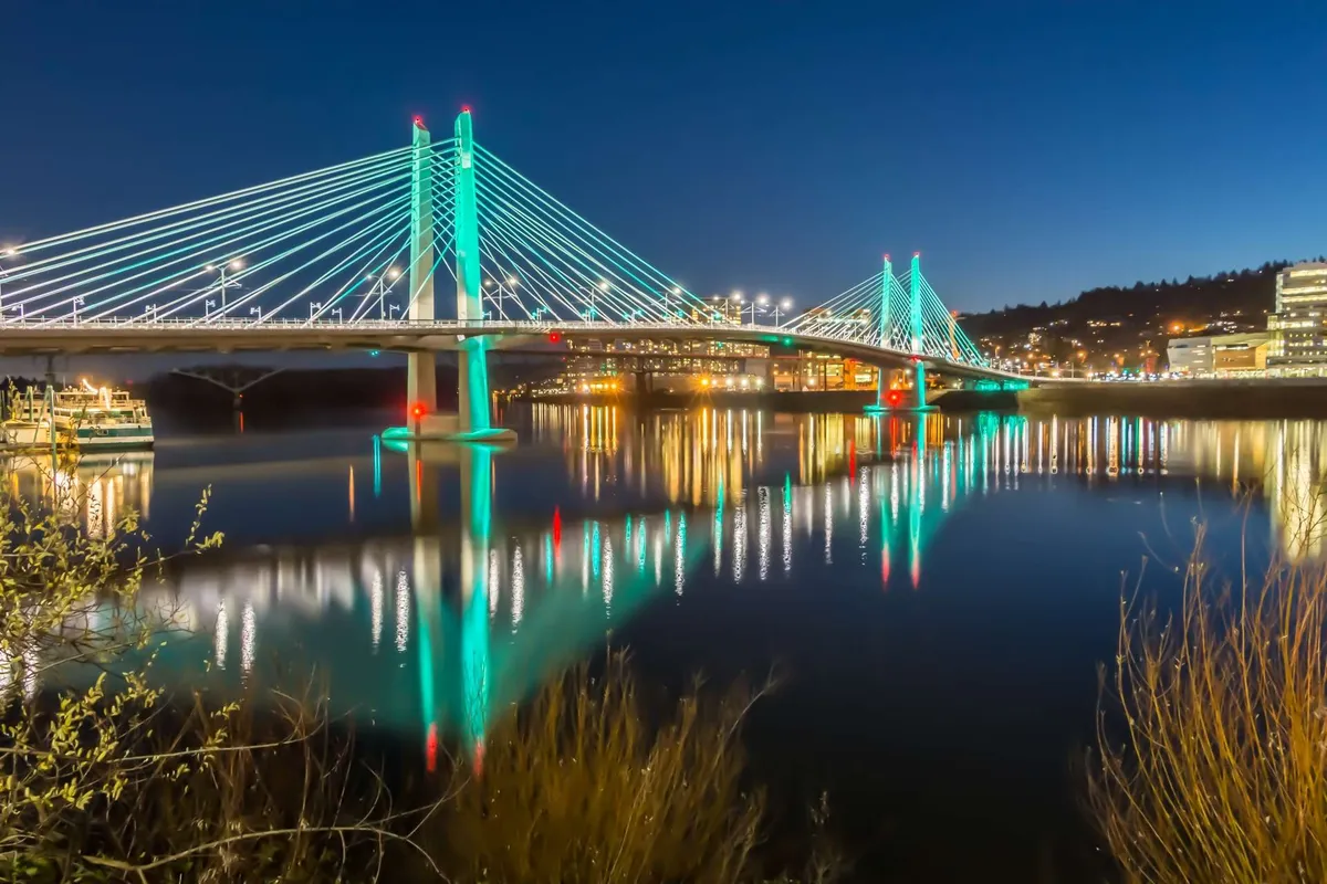 An illuminated city bridge arches over tranquil water, with city lights reflecting on the river and the urban skyline just beyond. The scene captures lively evening energy, suggesting opportunities for night walks, city exploration, and riverside activities.