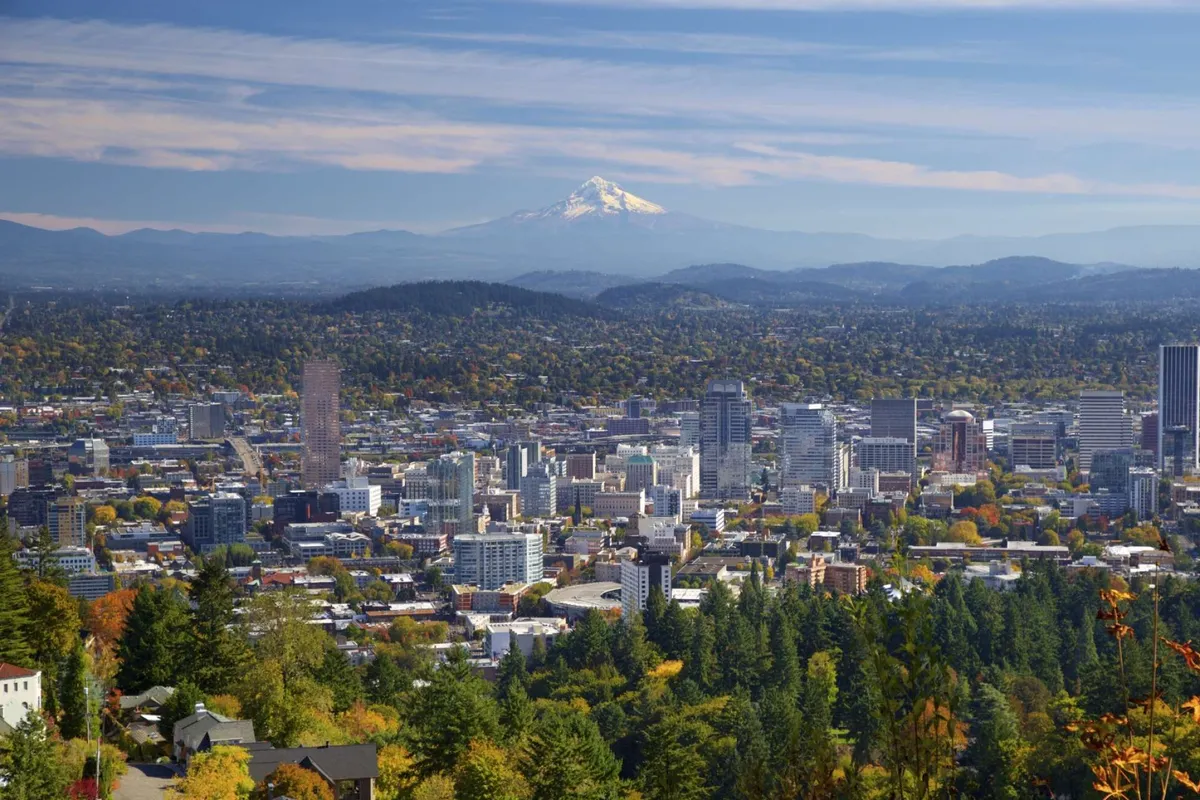This image captures a sweeping view of downtown Portland with Mount Hood rising in the distance, surrounded by vibrant autumn foliage. The city skyline is framed by the changing colors of trees, presenting a harmonious blend of urban life and natural beauty. The scene reflects an inviting atmosphere, ideal for family adventures and leisurely exploration.