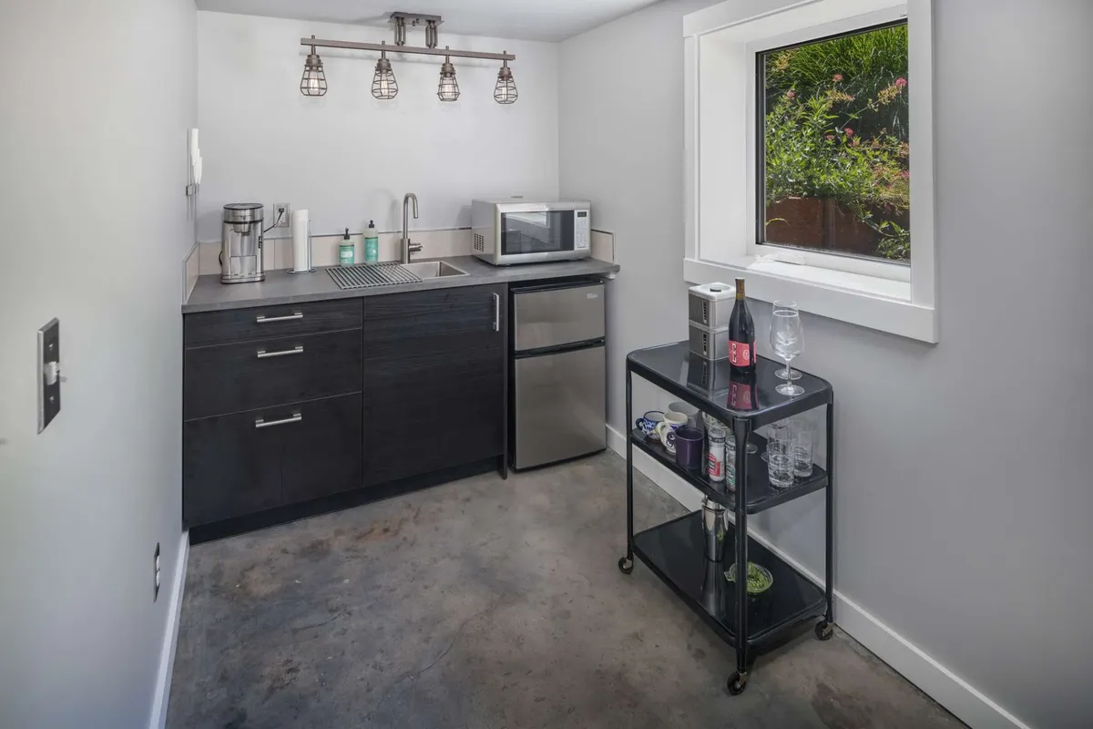 The image captures an interior view centered on sleek kitchen cabinetry, stainless steel appliances, and a window framing lush greenery. A bar cart adds a touch of hospitality, and the overall environment feels welcoming and homey.