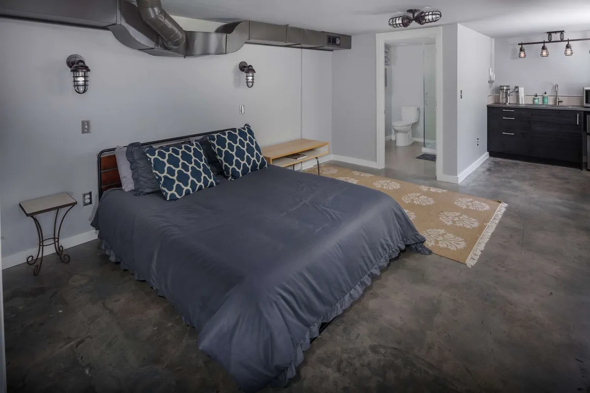 This image shows an inviting and comfortable bedroom area featuring a queen bed with decorative pillows, a small side table, and a glimpse of a kitchenette. The scene evokes a sense of relaxation and convenience, ideal for restful nights or enjoying breakfast in bed.